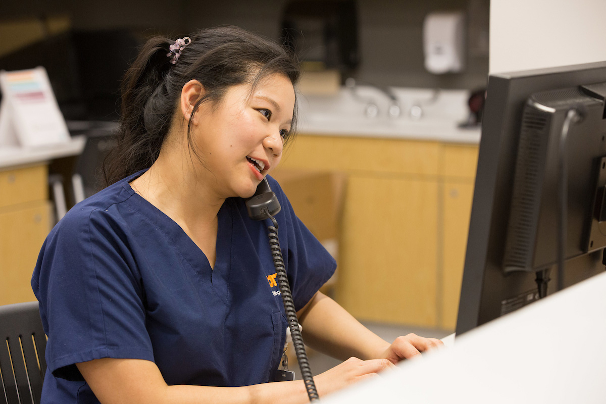 Student in scrubs on phone typing on a cumputer
